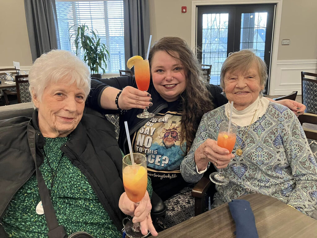 Two senior resident ladies and an employee are enjoying happy hour at a table, smiling and sipping their drinks.