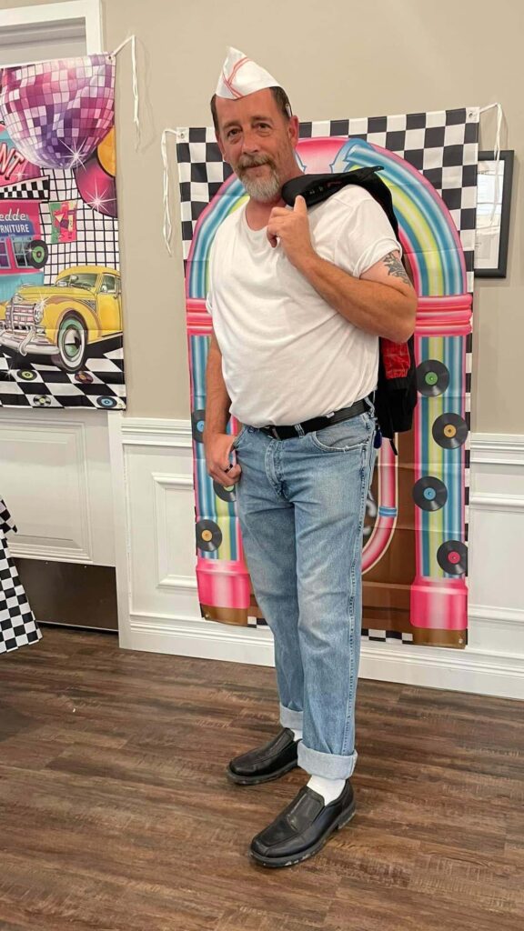 A man in a white shirt and jeans standing in front of a wall at Vitalia North Royalton senior living Retro happy hour event.