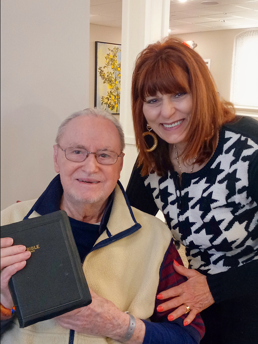 Senior man holding a Bible and smiling, sitting next to a woman with red hair who is also smiling, both in a senior living community.