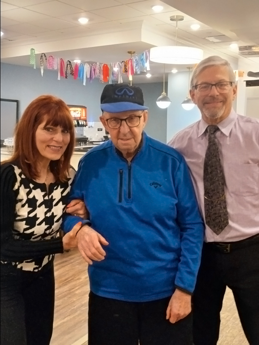 Senior man in a blue jacket and cap standing with a smiling woman in a black and white sweater and a man in a light purple shirt and tie at a Bible study in a senior living community.