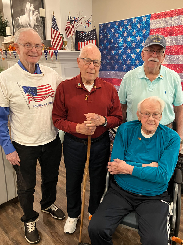 Four senior male veterans pose together in front of an American flag backdrop. They are smiling and dressed in patriotic clothing, with one using a walking cane.