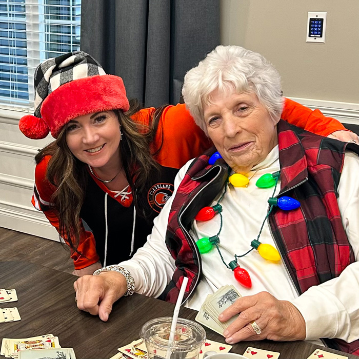 A senior resident and a staff member pose together while playing cards. The resident is wearing holiday lights around her neck, and the staff member wears a festive hat and jersey.