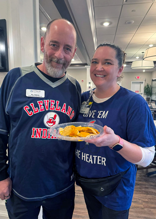 Two senior living team members smile together while holding a plate of nachos during a community game day.