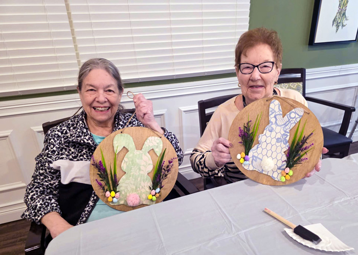 Two senior residents proudly hold up their handmade spring bunny crafts decorated with flowers and ribbon.