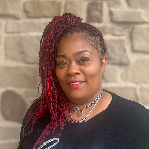 Kimshaya Cooper, Director of Wellness at Vitalia North Royalton, smiling in front of a stone wall, wearing a black top and silver jewelry with red braided hair.