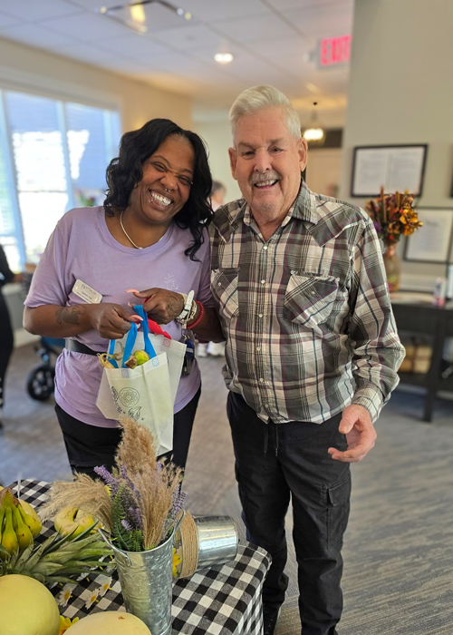 A female team member and a senior resident smile together at a table, standing beside a cheerful display of fresh produce and flowers at a farmers market event.