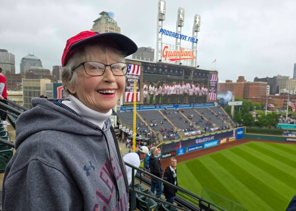 A senior woman smiles in the stands at Progressive Field with the Cleveland Guardians scoreboard and city skyline in the background on a cloudy game day.