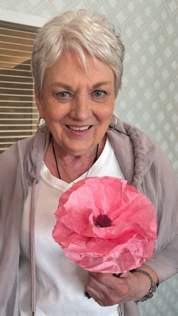 Smiling senior woman holding a large pink tissue paper flower during a creative crafting session.