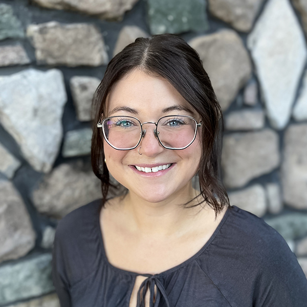 Jessie Cooper, Operations Coordinator at Vitalia North Royalton, smiling while wearing glasses and a charcoal blouse, standing in front of a stone wall.