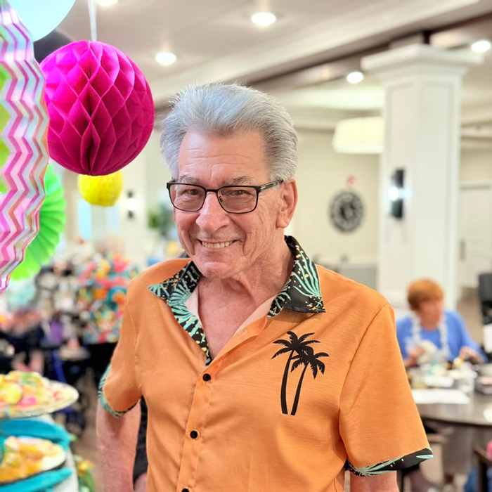 Senior man wearing a tropical palm tree shirt and smiling at a festive luau-themed party with colorful decorations.