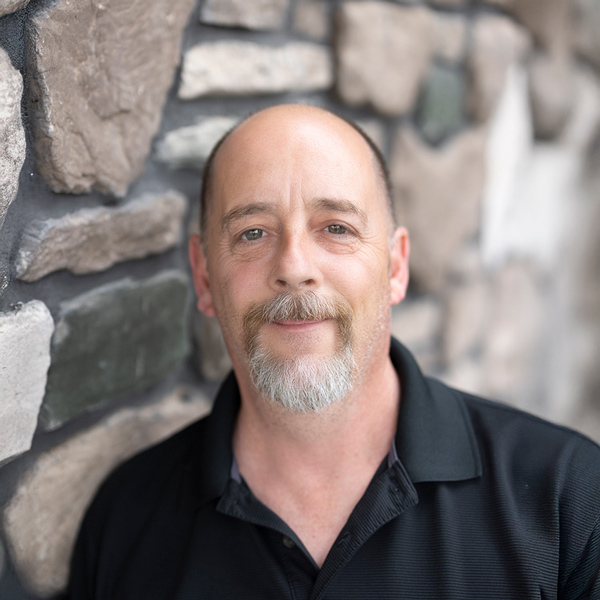 William Gibbons, Plant Operations Director at Vitalia North Royalton, wearing a black collared shirt and standing in front of a stone wall, smiling gently.