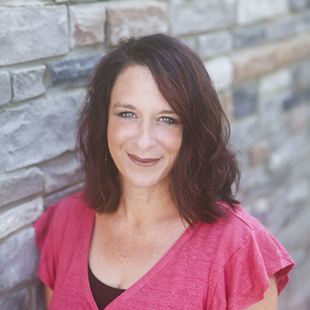 Julie Sabo, Executive Director at Vitalia North Royalton, wearing a deep pink top with flutter sleeves, standing against a stone wall background and smiling warmly.