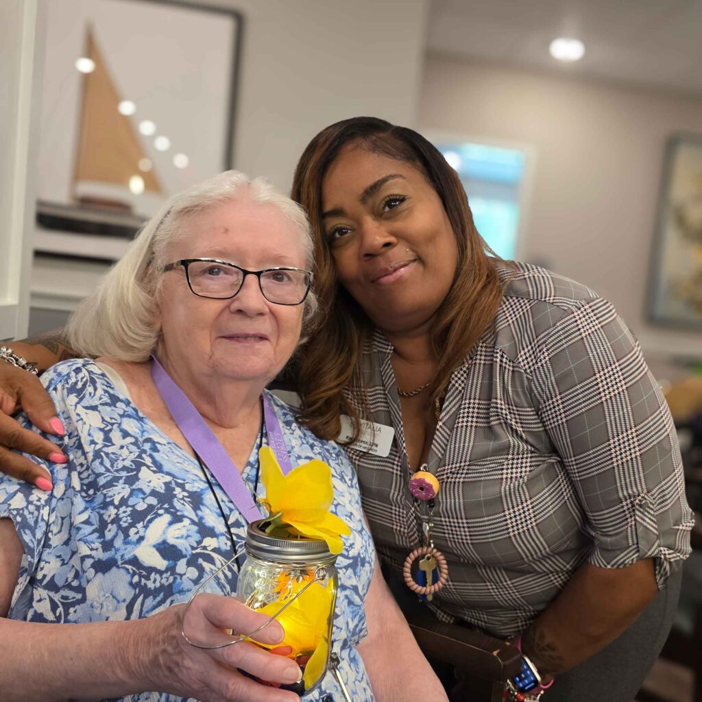 A senior woman proudly holds a decorated lantern craft while smiling beside a supportive team member, highlighting creativity and companionship in the community.