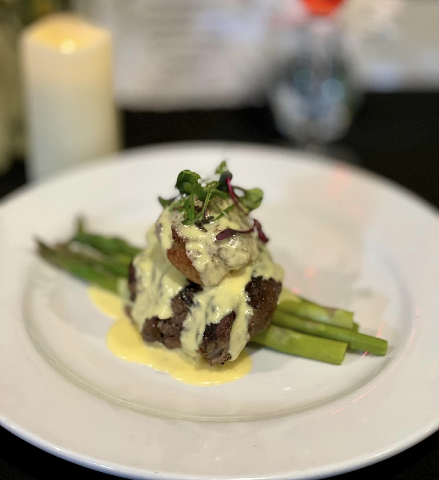 Plated gourmet steak with asparagus, topped with creamy hollandaise sauce and garnished with microgreens, set against a blurred background of a candle, flowers, and drink.