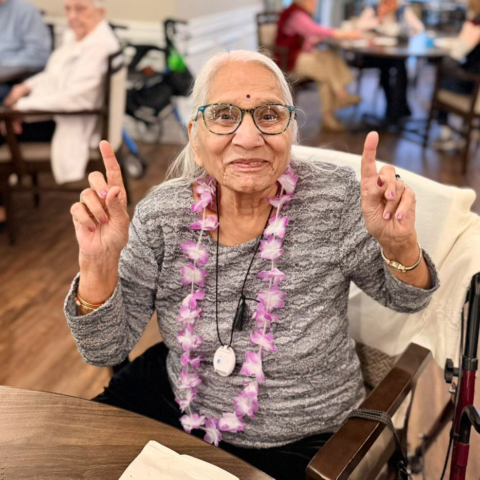 A senior woman at Vitalia North Royalton gives two "number one" signs, wearing a purple flower lei during a themed community event.