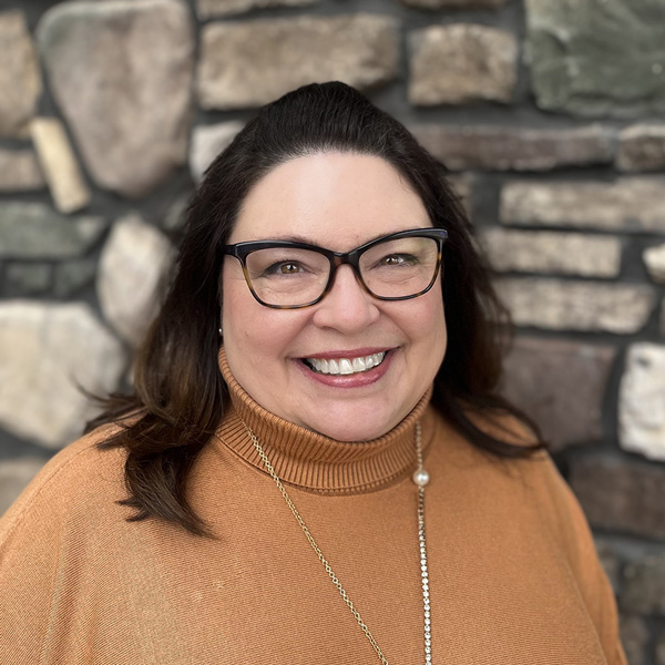 Melissa Reagon, Operations Coordinator at Vitalia North Royalton, smiling in front of a stone wall, wearing a tan turtleneck sweater, black glasses, and a long gold necklace.