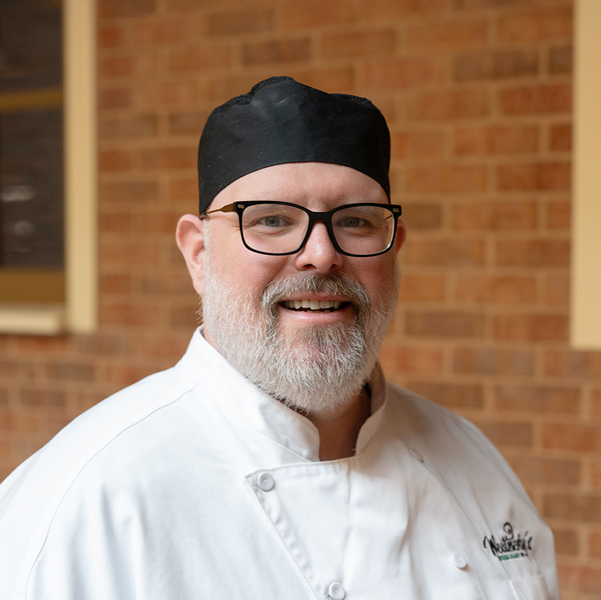 Andrew “Andy” Shelley, Culinary Director at Vitalia North Royalton, smiling in a professional headshot, wearing a white chef coat, black chef hat, and glasses, with a softly blurred brick background.