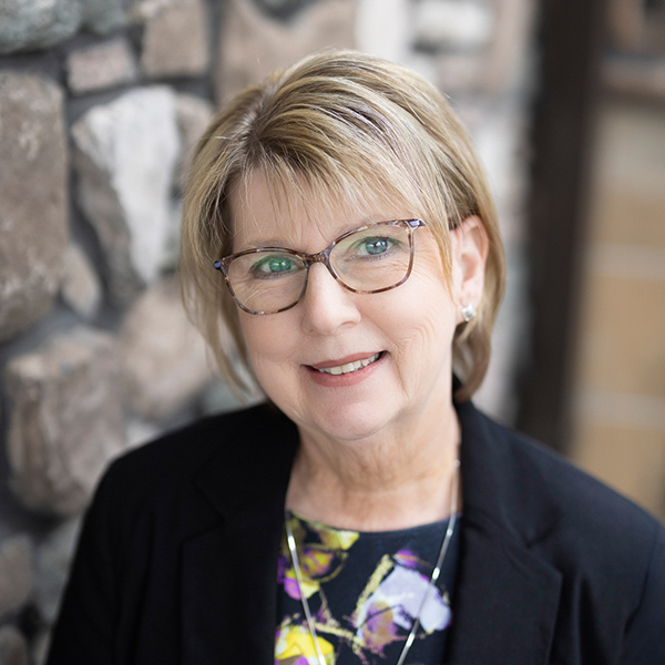 Lori Robinson, Senior Living Coordinator at Vitalia North Royalton, smiling in a professional headshot, wearing glasses, a black blazer, and a floral blouse, with a softly blurred stone wall background.