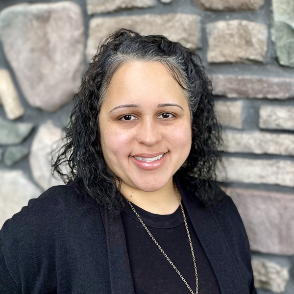 Marisol Amill, Memory Care Director at Vitalia North Royalton, smiling in a professional headshot, wearing a black top and necklace, with a softly blurred stone wall background.