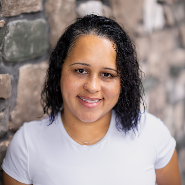 Marisol Amill, Memory Care Director at Vitalia North Royalton, smiling in a professional headshot with short curly dark hair, a white top, and a softly blurred stone wall background.