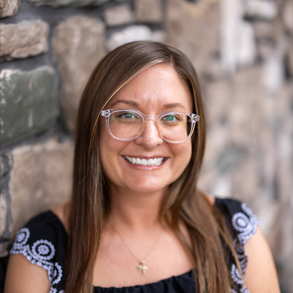 Melanie Knautz, Wellness Director at Vitalia North Royalton, smiling in a professional headshot with long straight highlighted hair, clear glasses, a black top with embroidered sleeves, and a softly blurred stone wall background.