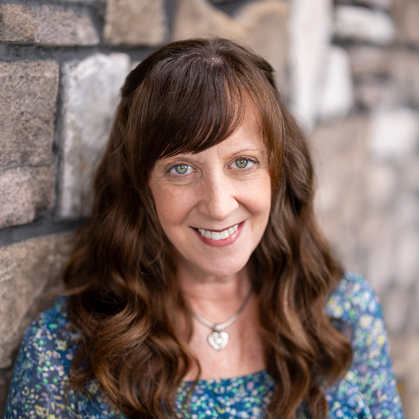 Michaela Johnson, Operations Coordinator at Vitalia North Royalton, smiling in a professional headshot with long wavy brown hair, a floral blue top, a heart pendant necklace, and a softly blurred stone wall background.